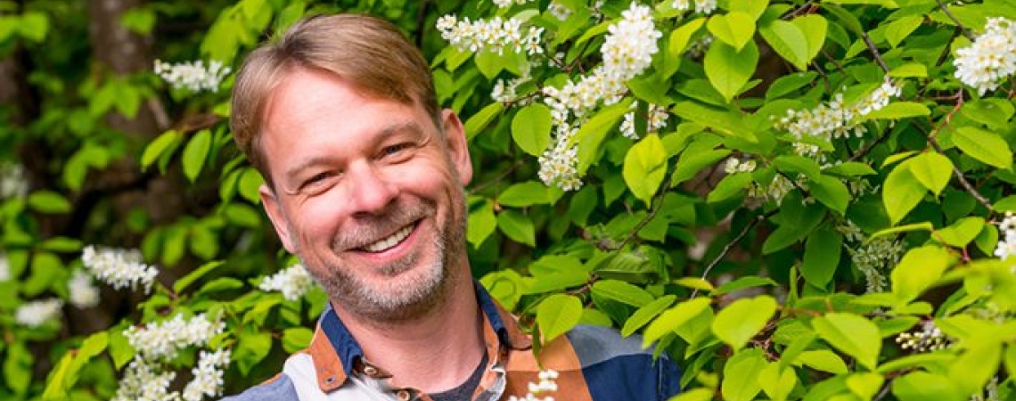 Mann mit kariertem Hemd und Hut steht lächelnd vor blühendem Strauch.