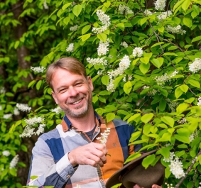 Mann mit kariertem Hemd und Hut steht lächelnd vor blühendem Strauch.