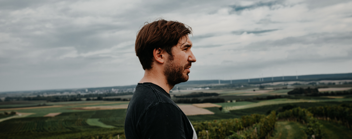 Mann im Profil vor weitläufiger Landschaft mit Wiesen und bewölktem Himmel.