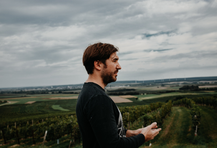 Mann im Profil vor weitläufiger Landschaft mit Wiesen und bewölktem Himmel.