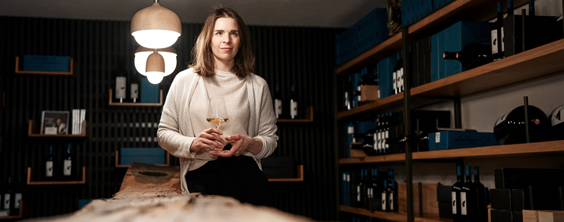 Frau mit Weinglas in modernem Weinkeller mit hölzernen Regalen voller Flaschen.
