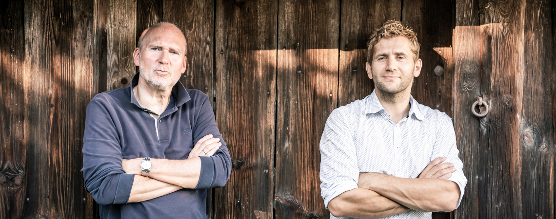Zwei Männer lehnen mit verschränkten Armen an einer Holzwand.