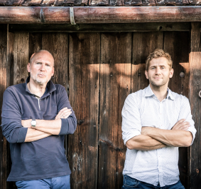 Zwei Männer lehnen mit verschränkten Armen an einer Holzwand.