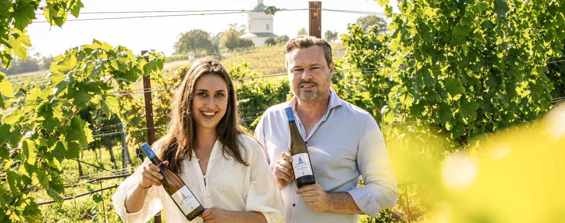 Zwei Personen im Weinberg halten Weinflaschen, umgeben von grünen Reben.