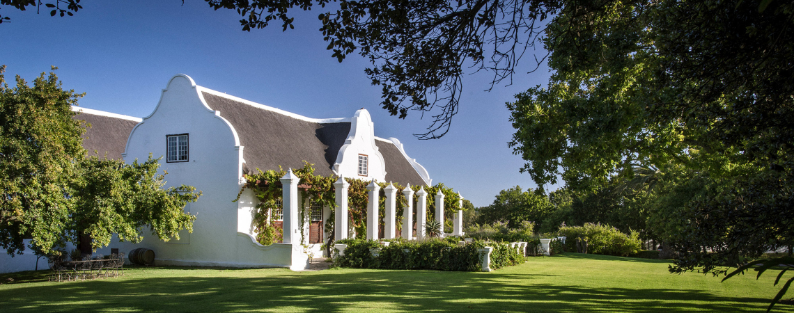 Weißes, traditionelles Gebäude mit Giebeldach, umgeben von gepflegtem Rasen und Bäumen.