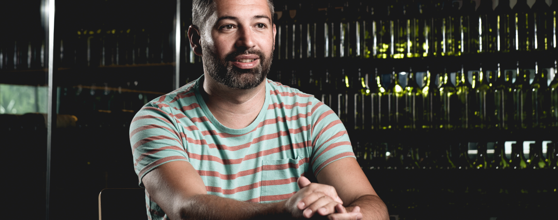 Mann in gestreiftem T-Shirt sitzt vor einem Regal mit Weinflaschen.