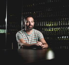 Mann in gestreiftem T-Shirt sitzt vor einem Regal mit Weinflaschen.