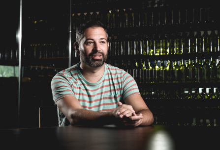 Mann in gestreiftem T-Shirt sitzt vor einem Regal mit Weinflaschen.