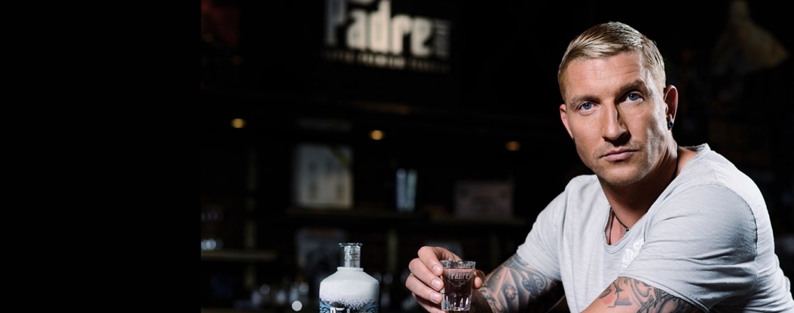 Mann mit Tattoos hält Getränk an einer Bar mit Flasche im Hintergrund.