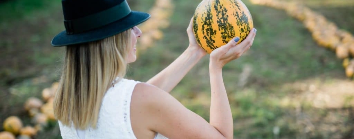 Person im weißen Kleid, schwarzem Hut, hält gestreiften Kürbis auf Feld.