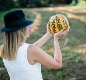 Person im weißen Kleid, schwarzem Hut, hält gestreiften Kürbis auf Feld.