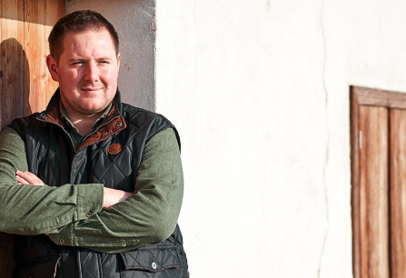 Mann in schwarzer Weste und grünem Hemd lehnt an Holztür, draußen bei Sonnenlicht.