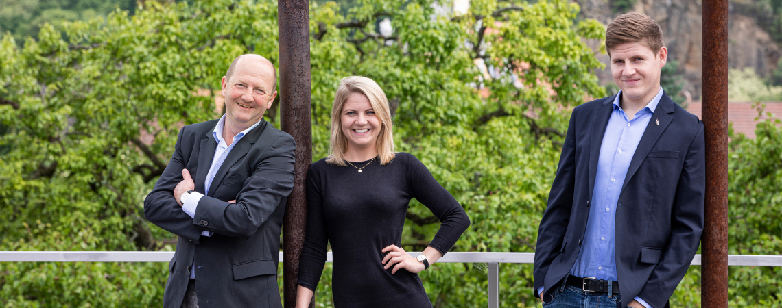 Drei Personen posieren lächelnd vor grünem Wald und Kirchturm im Hintergrund.