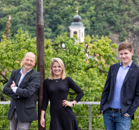 Drei Personen posieren lächelnd vor grünem Wald und Kirchturm im Hintergrund.