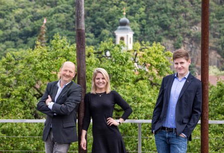 Drei Personen posieren lächelnd vor grünem Wald und Kirchturm im Hintergrund.