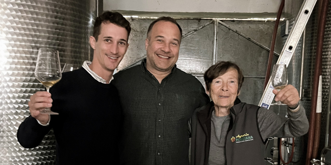 Drei Personen mit Weingläsern stehen vor edelstahlfarbenen Industriebehältern.