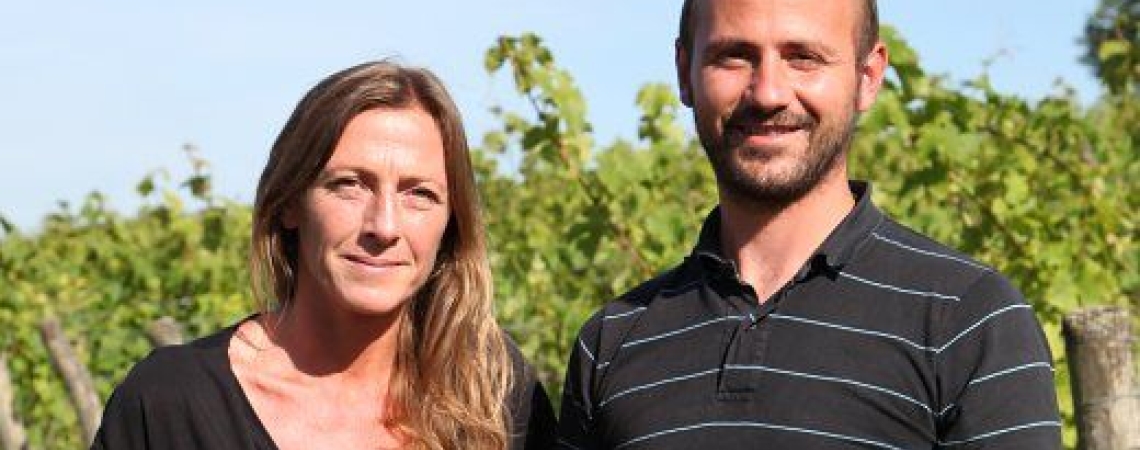 Zwei Personen stehen lächelnd in einem sonnigen Weinberg.