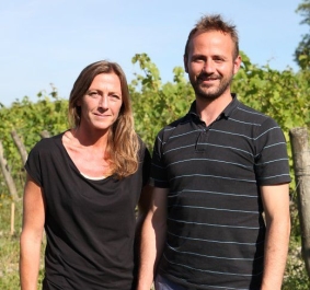 Zwei Personen stehen lächelnd in einem sonnigen Weinberg.