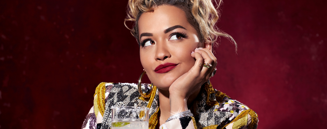 Person mit lockigem Haar und gemusterter Jacke hält ein Getränk vor rotem Hintergrund.