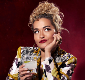 Person mit lockigem Haar und gemusterter Jacke hält ein Getränk vor rotem Hintergrund.