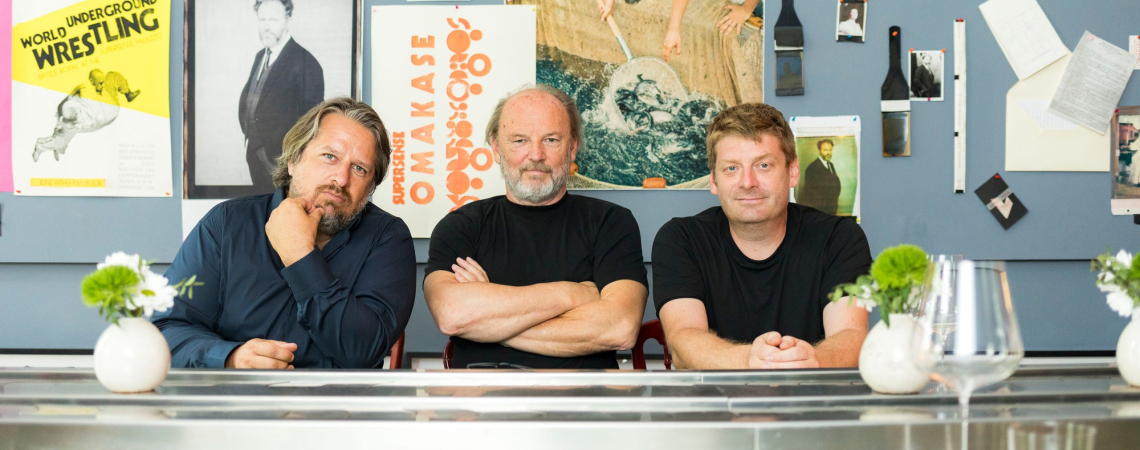 Drei Männer sitzen an einem Tisch mit bunter Posterwand im Hintergrund.