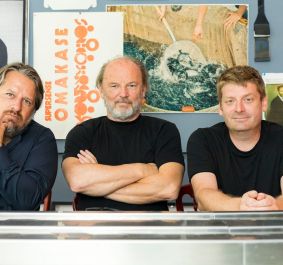 Drei Männer sitzen an einem Tisch mit bunter Posterwand im Hintergrund.