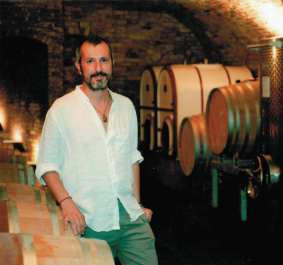 Mann in weißem Hemd steht in einem stimmungsvollen Weinkeller vor Fässern.