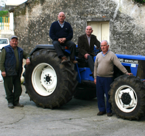 Fünf Männer posieren neben und auf einem blauen Traktor vor rustikaler Wand.