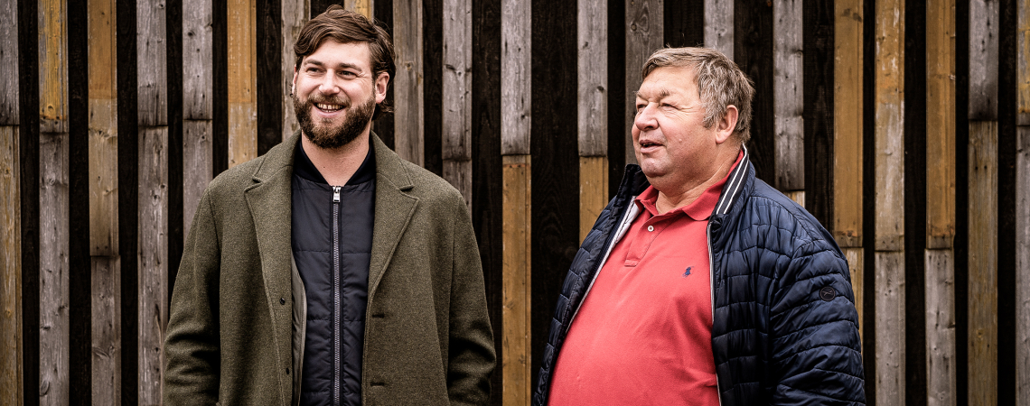 Zwei Männer in Mänteln stehen vor einer Holzwand und lächeln.