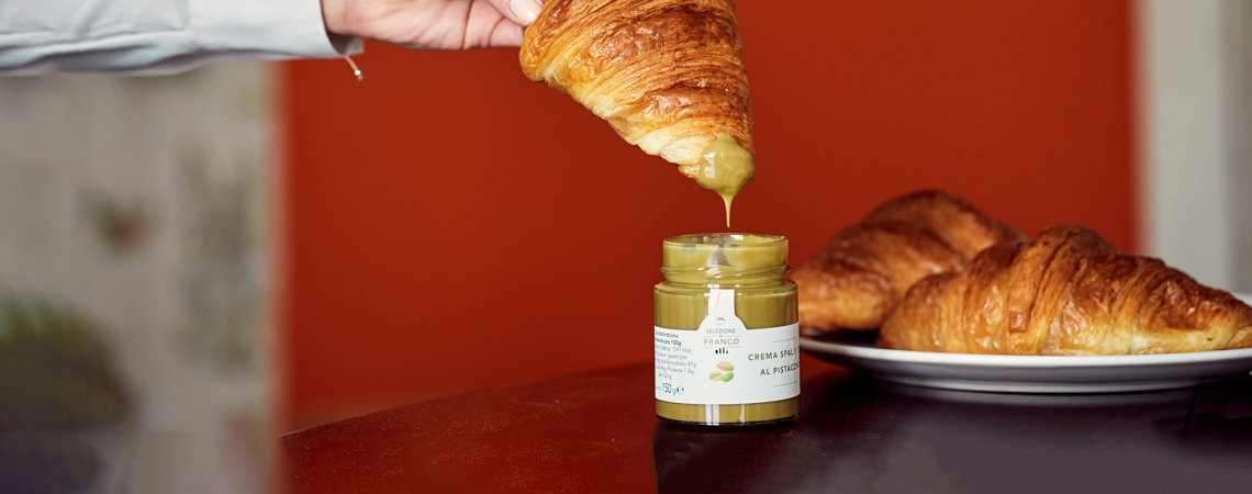Croissant wird in Pistaziencreme getunkt, Hintergrund orange, daneben ein Teller mit Croissants.