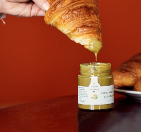 Croissant wird in Pistaziencreme getunkt, Hintergrund orange, daneben ein Teller mit Croissants.