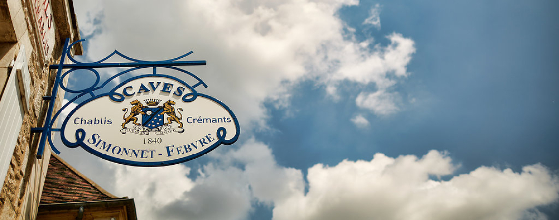 Blaues Schild mit Wappen vor einem Gebäude, bewölkter Himmel im Hintergrund.