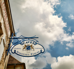 Blaues Schild mit Wappen vor einem Gebäude, bewölkter Himmel im Hintergrund.