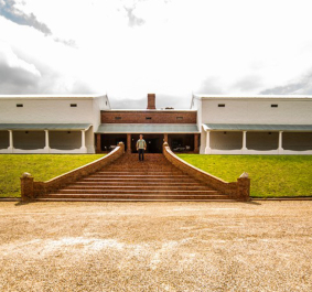 Weißes Gebäude mit breiter Treppe auf einer grünen Rasenfläche, unter dramatischem Himmel.