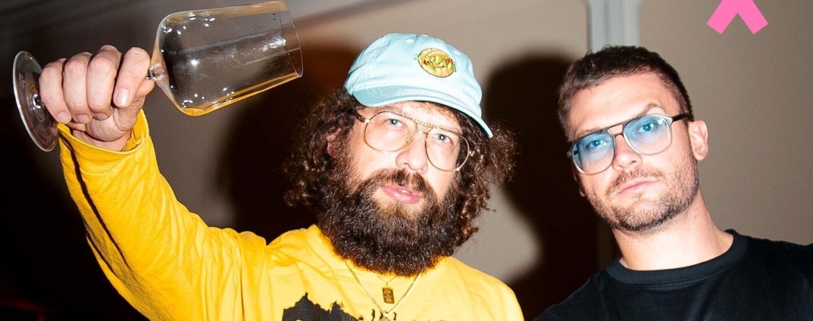 Zwei Männer mit Brillen, einer hält ein Weinglas, vor dunklem Hintergrund.