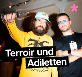 Zwei Männer mit Brillen, einer hält ein Weinglas, vor dunklem Hintergrund.