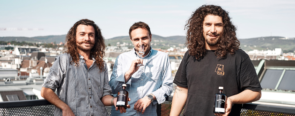 Drei Männer auf Balkon, zwei halten Flaschen mit dunklem Etikett vor Stadtpanorama.