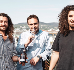 Drei Männer auf Balkon, zwei halten Flaschen mit dunklem Etikett vor Stadtpanorama.