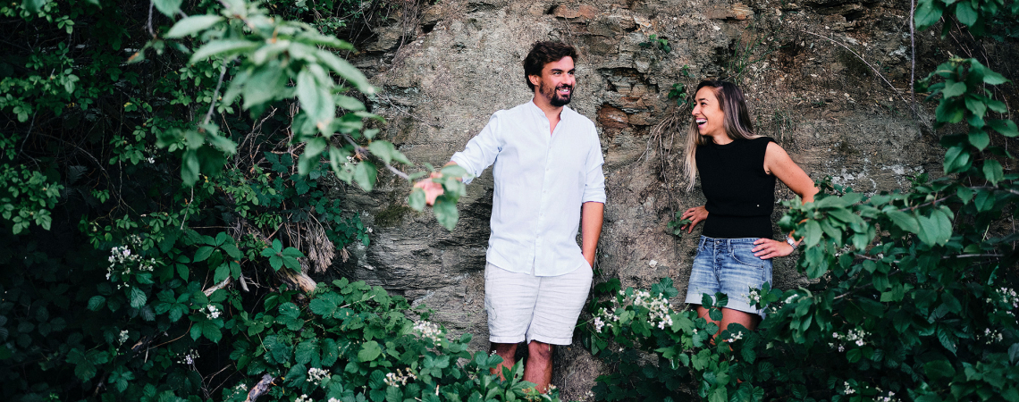 Zwei lachende Personen vor einer mit Pflanzen bewachsenen Steinmauer.