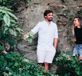 Zwei lachende Personen vor einer mit Pflanzen bewachsenen Steinmauer.
