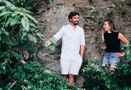 Zwei lachende Personen vor einer mit Pflanzen bewachsenen Steinmauer.