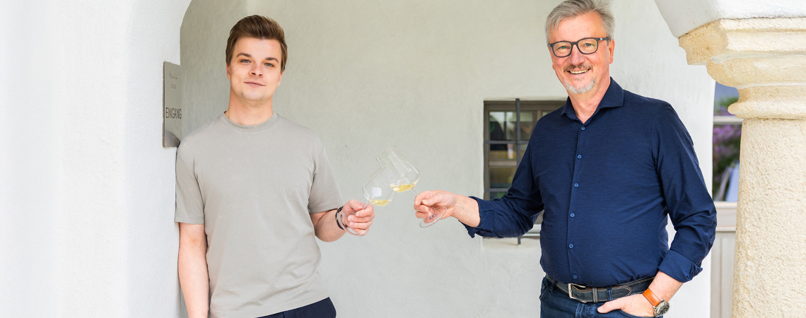 Zwei Männer stoßen mit Weißweingläsern vor hellem Hintergrund an.
