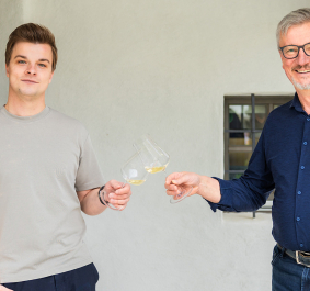 Zwei Männer stoßen mit Weißweingläsern vor hellem Hintergrund an.