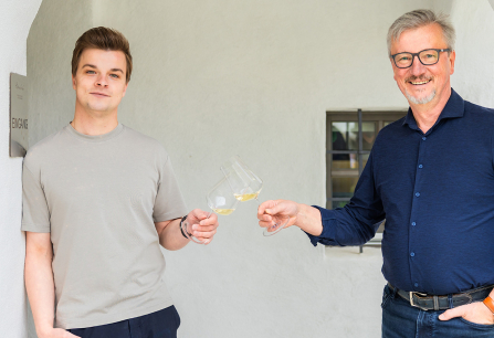 Zwei Männer stoßen mit Weißweingläsern vor hellem Hintergrund an.