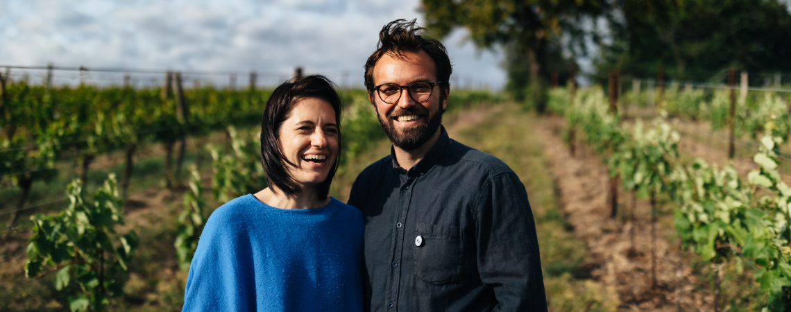 Paar lächelt in einem sonnigen Weinberg mit grünem Laub.
