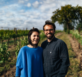 Paar lächelt in einem sonnigen Weinberg mit grünem Laub.