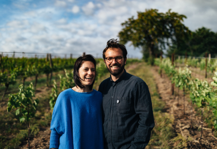 Paar lächelt in einem sonnigen Weinberg mit grünem Laub.