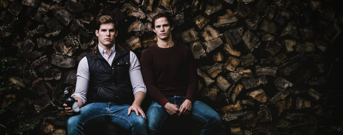 Zwei Männer vor gestapeltem Holz, lässig sitzend, in Freizeitkleidung.