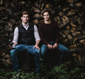 Zwei Männer vor gestapeltem Holz, lässig sitzend, in Freizeitkleidung.