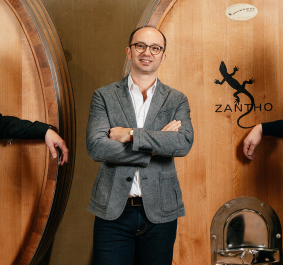 Drei Männer posieren lächelnd vor großen Holzfässern in einer Weinkellerei.